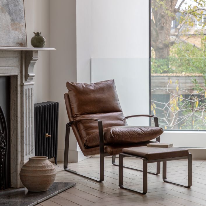 The Gallery Direct Fabien Footstool has a cushioned top upholstered in Brown Leather, with a dark metal frame. A matching armchair is also available