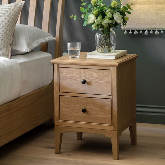 Gallery Direct Malvern 2 Drawer Bedside Cabinet is constructed using warm oak solids and veneers with natural grain patterns, complemented by dark metal handles and with slim tapered legs